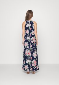 Kvinna som står med ryggen mot oss, iförd en marinblå blommig ärmlös maxiklänning med rosa och vita blommor samt ljusrosa högklackade skor.