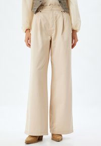 Beige wijde broek van zachte katoen met plooien aan de voorkant, twee zijzakken en twee knoopaccenten op de tailleband.