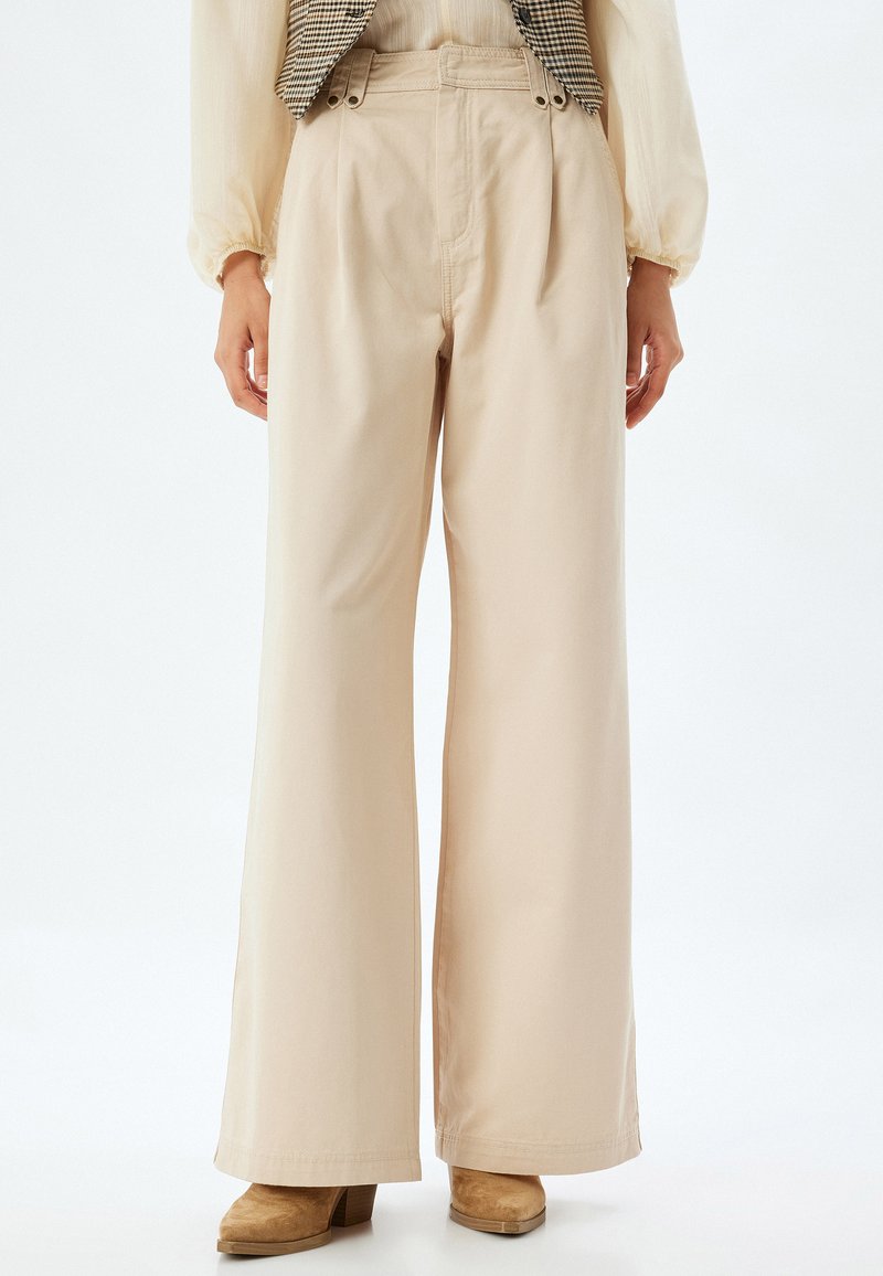 Beige wijde broek van zachte katoen met plooien aan de voorkant, twee zijzakken en twee knoopaccenten op de tailleband.