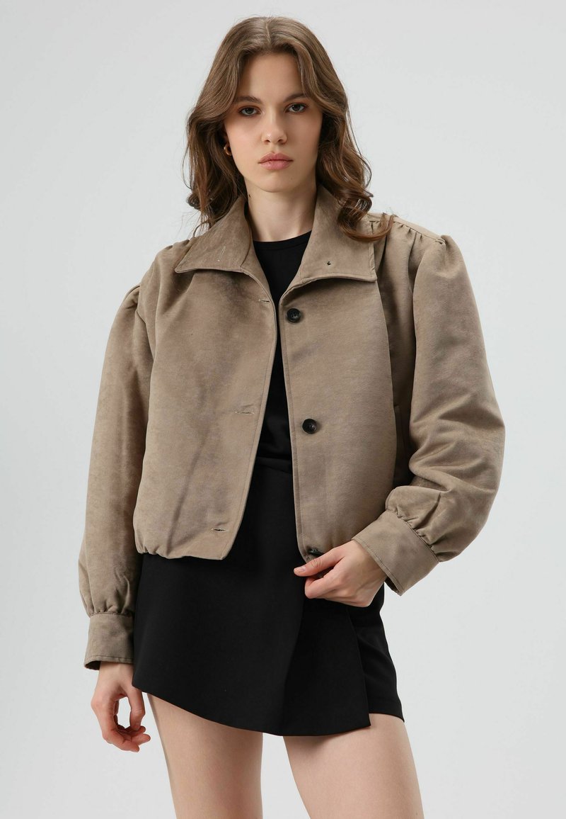 Light brown cropped jacket in soft fabric with a pointed collar, button closure, and puffy sleeves, worn over a black dress.
