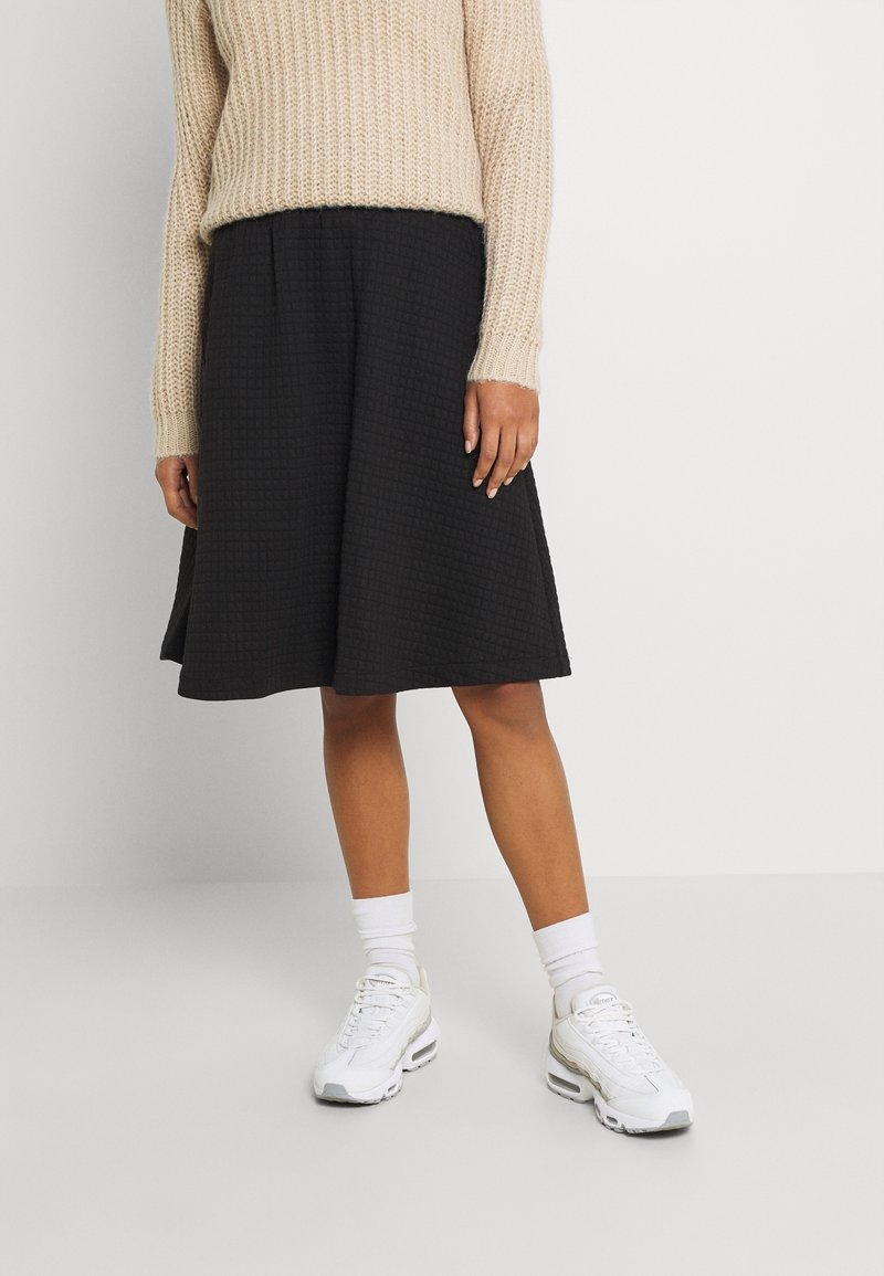 Černá, strukturovaná sukně ve tvaru A s vaflovým vzorem. V kombinaci s béžovým pleteným svetrem a bílými teniskami, doplněné bílými ponožkami.