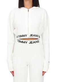 Bílá pletená zkrácená mikina s kapucí a předním zipem, s širokým páskem vyšitým nápisem "TOMMY JEANS" v černé barvě a dlouhými rukávy.