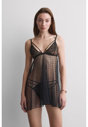 Frau trägt transparente schwarze Spitzen-Nachtwäsche mit Polka-Dots, steht vor einer schlichten hellgrauen Wand und schaut nach vorne.