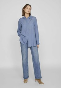 Modra srajca s dolgimi rokavi, kombinirana z svetlo modrimi kavbojkami in bež sistematičnimi čevlji s konicami. Gladek material in sproščen kroj.