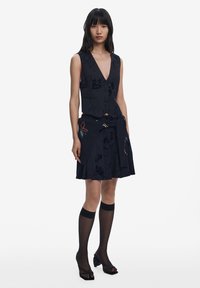 Svart ärmlös topp med blommönster, i kombination med en svart plisserad kjol. Outfits kompletteras av knähöga svarta strumpor och högklackade sandaler.