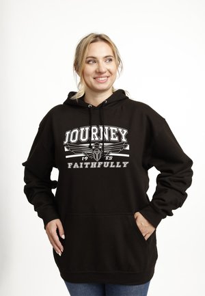 Donna sorridente con capelli biondi che indossa un cappuccio nero con il testo e la grafica "JOURNEY 1983 FAITHFULLY", mani nelle tasche, su sfondo bianco.