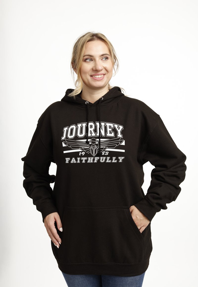 Donna sorridente con capelli biondi che indossa un cappuccio nero con il testo e la grafica "JOURNEY 1983 FAITHFULLY", mani nelle tasche, su sfondo bianco.