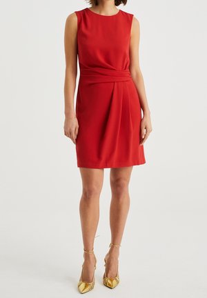 Femme portant une robe rouge sans manches mi-longue avec un détail plissé à la taille et des talons hauts dorés à brides à la cheville, sur fond blanc.