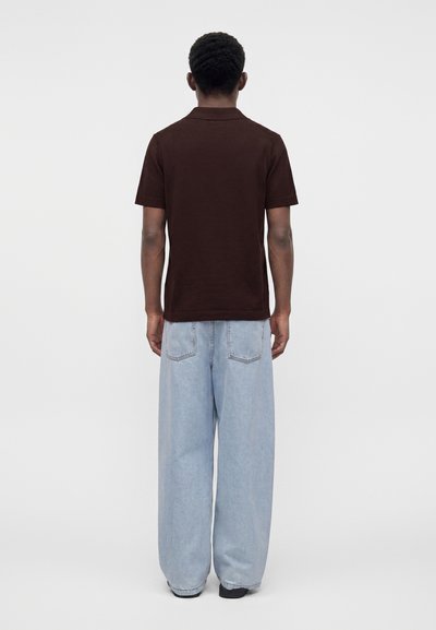 Homme debout de dos, portant une chemise marron foncé à manches courtes et un jean large bleu clair sur un fond uni.