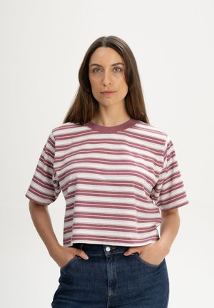 Vrouw met lang bruin haar, gekleed in een cropped wit shirt met korte mouwen en bordeauxrode strepen, en donkerblauwe spijkerbroek, met handen in de zakken.