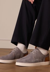 Sneaker in suede grigio con dettagli in pelle marrone chiaro, lacci bianchi e suola bianca spessa; abbinati a calzini bianchi e pantaloni scuri.