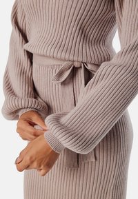 Geribd gebreide jurk in zacht beige, met een fitted taille en een strikceintuur, lange mouwen met brede manchetten en een textuurdetail in de stof.