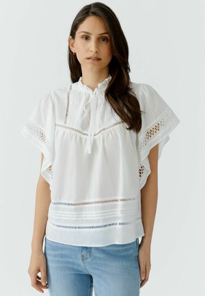 Vrouw met lang donker haar die een witte blouse draagt met kanten details en geplooide mouwen, gecombineerd met lichtblauwe spijkerbroek tegen een effen achtergrond.