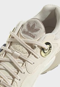 Krämfärgad sneaker med texturerad mesh-tunga, guldfärgade detaljer och platta snören. Har perforeringar och en rundad form för komfort.