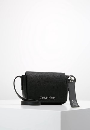 Sac bandoulière en cuir noir texturé avec logo Calvin Klein en argent et étiquette en cuir de marque attachée sur une surface blanche.