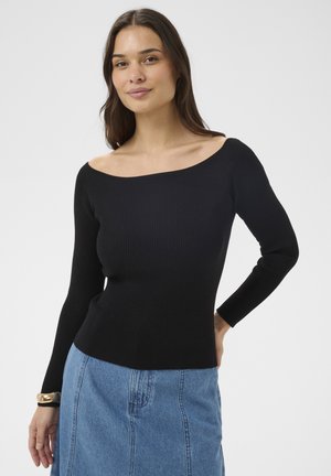 Mlada ženska z dolgimi temnimi lasmi, oblečena v črn rebrast pulover brez ramen in modro jeans krilo, na belem ozadju.