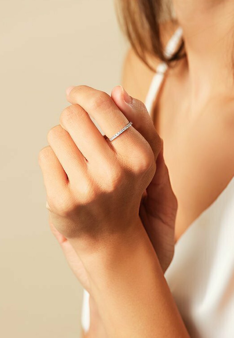 Silberner Ring mit einer Reihe kleiner klarer Steine entlang des Bands, getragen an einer Hand mit leicht gebräunter Haut vor einem neutralen Hintergrund.