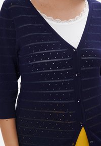 Cardigan blu navy con scollo a V, maniche a metà e un motivo di strisce orizzontali e pois. Presenta bottoni e un top bianco a strati sotto.