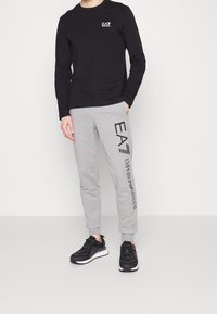 Černé tričko s dlouhým rukávem s logem EA7; šedé tepláky s textem „EA7 Emporio Armani“ a černé sportovní boty s b ílými podrážkami.