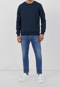 Sudadera de cuello redondo azul marino con un logotipo en relieve, combinada con jeans de color azul claro y zapatillas blancas, de pie frente a un fondo neutro.