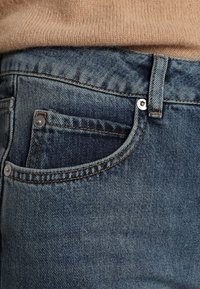 Modré denimové džíny mají přední kapsu se stříbrným knoflíkem a kontrastním oranžovým šitím. Látka vykazuje mírnou strukturu a vyblednutí.