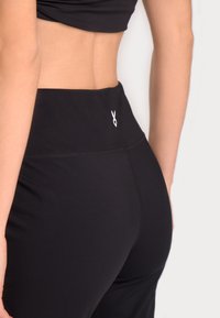 Črne atletske tajice z visokim pasom, opremljene s sploščenimi šivi in majhnim belim logotipom na hrbtni strani za dodaten poudarek.