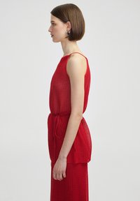 Vestido de punto rojo con un corte holgado, cinturilla ajustable y tirantes finos. Presenta una superficie texturada y elementos de diseño en capas.