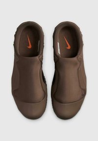 Zapatos tipo slip-on marrones con un material superior suave y flexible. Presenta acentos laterales negros y un pequeño logo naranja en la plantilla.