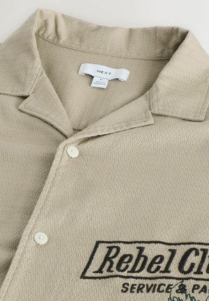 Beežika tooniga tekstuuriga nööbitav särk kraega, millel on kaks valget nööpi ja musta tikandiga tekst "Rebel Club Service & Part" esiküljel.