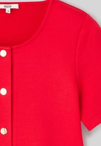 Haut rouge à manches courtes avec un col rond, cinq boutons blancs sur le devant, et un tissu en coton lisse avec une légère brillance.