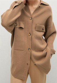 Camicetta beige in maglia con bottoni, due tasche frontali, colletto tradizionale e tessuto testurizzato. Presenta bottoni marroni e una vestibilità oversize.