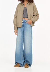 Béžová bomber bunda, šedý cropped top, světlé modré široké džíny a hnědé semišové boty. Hladký materiál, uvolněný střih a neformální styl.