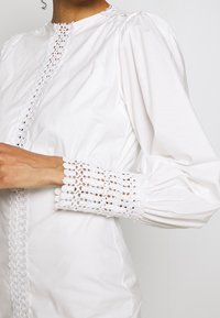 Gros plan d'une personne portant un chemisier blanc à manches longues avec une bordure en dentelle sur l'avant et des poignets en dentelle complexes aux manches.