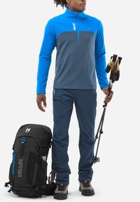 Blaues und graues Fleece-Pullover mit Viertelreißverschluss, marineblaue Hose, schwarzer Rucksack mit blauen Akzenten und Trekkingstöcke. Strukturierte Materialien, funktionales Design.