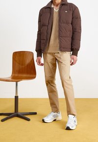 Brązowa puchowa kurtka, beżowy sweter dziany, beżowe spodnie i srebrne trampki. Drewniane krzesło obok; żółta podłoga dodaje kontrastu.