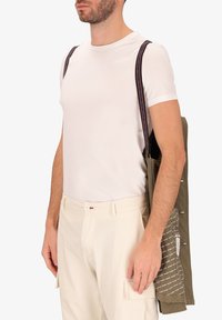 Giacca utilitaria verde con fodera a fantasia, indossata sopra una maglietta bianca, abbinata a pantaloni cargo color crema. Cinghie regolabili visibili.