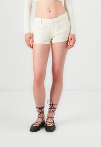 Shorts corti color crema con un design sartoriale, dotati di tasche frontali e una texture liscia. Abbinati a scarpe ballerine nere con lacci.