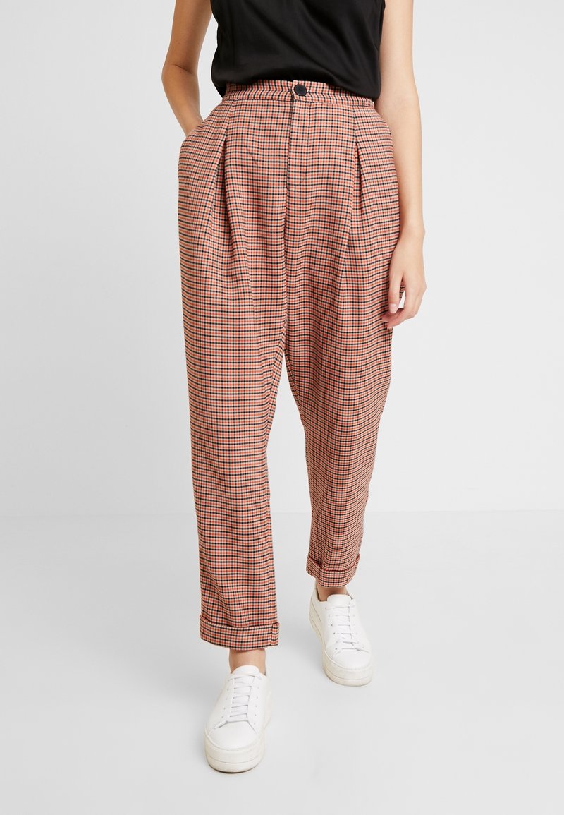 Femme portant un pantalon à carreaux rose et noir taille haute avec des plis et des baskets blanches, debout devant un fond clair uni.