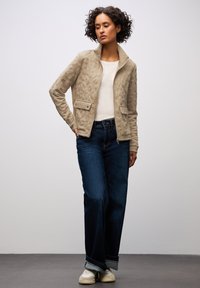 Beige vadderad jacka med ståkrage, två framfickor, baren över en vit topp, ihop med vida mörka jeans och vita sneakers.
