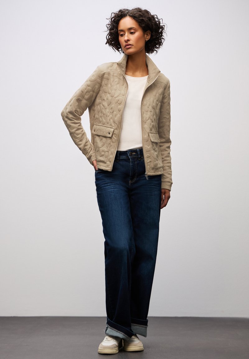 Beige vadderad jacka med ståkrage, två framfickor, baren över en vit topp, ihop med vida mörka jeans och vita sneakers.