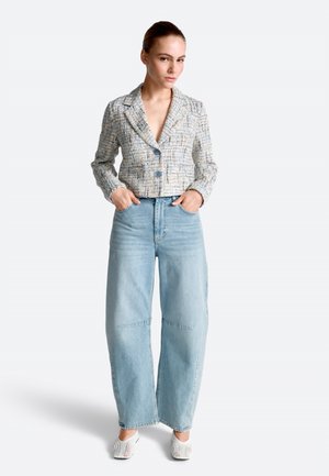 Femeie purtând un blazer scurt, texturat, cu nasturi mari, blugi albaștri cu picioare largi și pantofi albi slip-on din plasă, stând cu mâinile în buzunare.