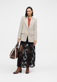 Grijze blazer over een roestkleurige top, gecombineerd met een zwarte kanten rok en bruine laarzen met hak. Een bruine tas met schapenvachtbies maakt de outfit compleet.