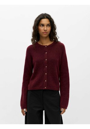 Vrouw draagt een donkerrode, met knopen gesloten, trui met lange mouwen en een zwarte broek, staand tegen een effen lichte achtergrond.