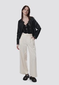 Giacca di pelle nera con borchie argentate, top nero aderente con dettagli floreali, pantaloni larghi color crema e sandali neri robusti.