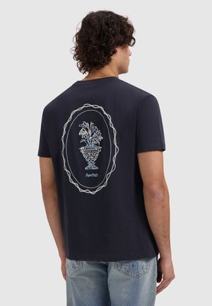 Persoon met krullend haar die een zwart T-shirt draagt met daarop geborduurde verwelkte bloemen in een decoratieve vaas, omlijst door een touwontwerp op de achterkant.