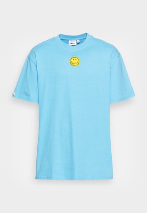 Lyseblå T-shirt med korte ærmer og et lille gult smilende ansigt placeret centreret under halsudskæringen på en ensfarvet baggrund.