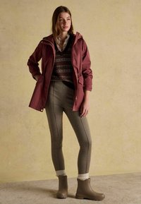 Burgundowa wodoodporna kurtka z kapturem, wzorzysty sweter pod spodem, dopasowane spodenki w kratę i brązowe botki za kostkę z teksturowaną podeszwą.