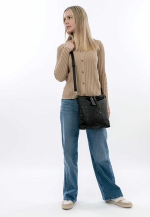 Bolso cruzado de cuero negro con textura suave, correa ajustable y un detalle con patrón. Modelo lleva un suéter beige de canalé y jeans.