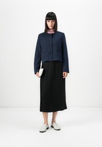 Femme avec des cheveux raides et foncés portant une veste bleu marine à boutons, un col roulé rose, une jupe plissée noire et des mocassins blancs, tenant une petite pochette blanche.