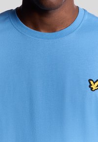 Ljust blå bomull t-shirt med rund hals och en svart och gul fågologotyp på vänster sida. Slät textur och avslappnad design.